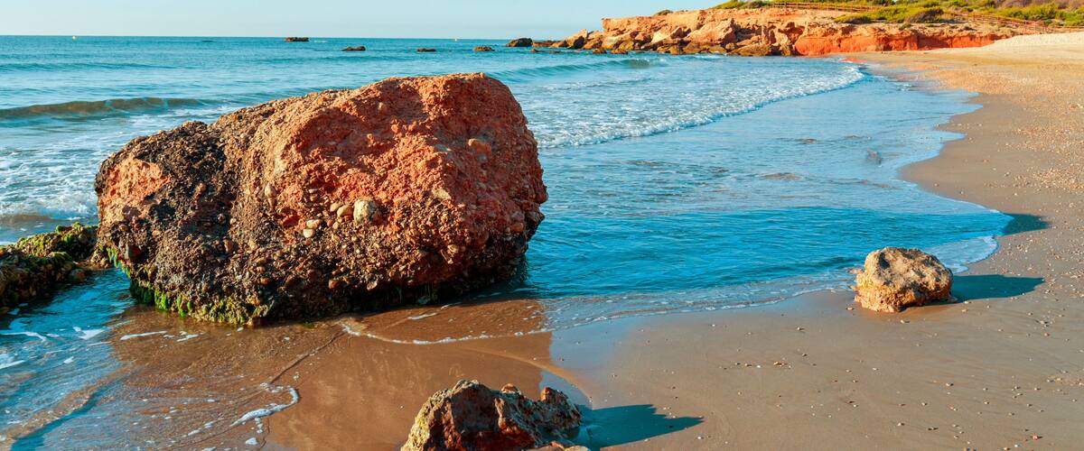 A beach with large rocks, calm water, and a clear sky.