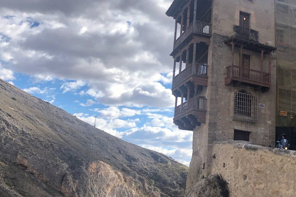 Un paisaje montañoso agreste con un edificio histórico que presenta balcones de madera y un muro de piedra.