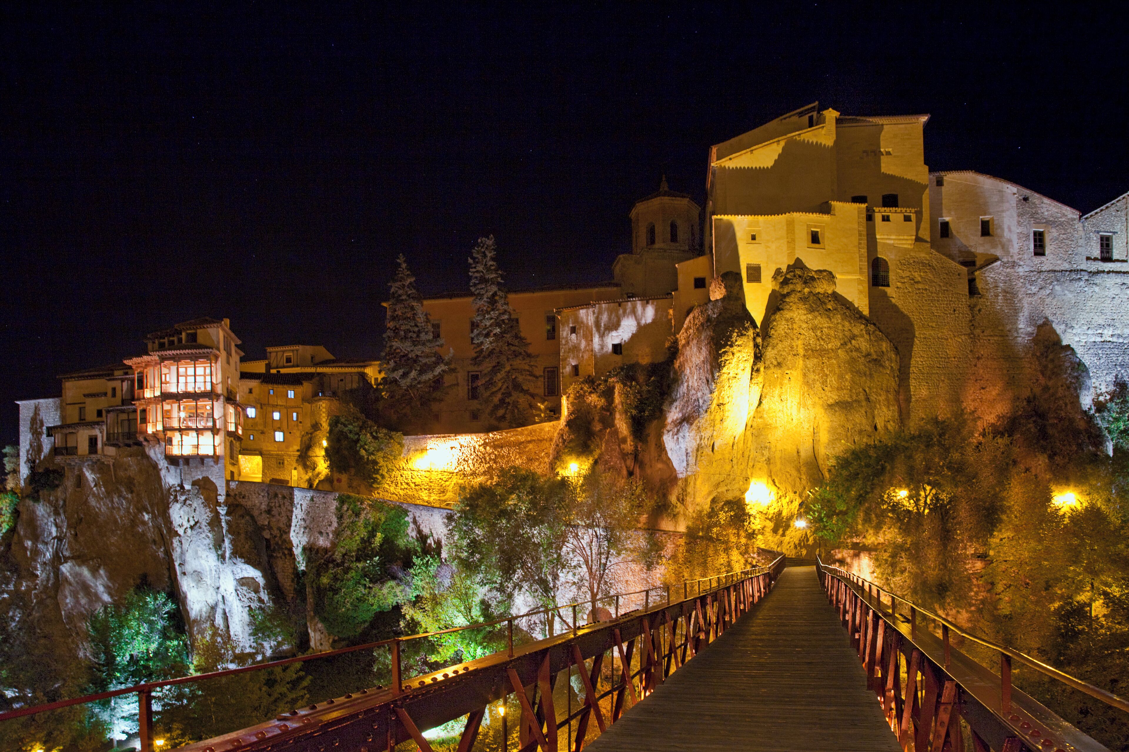A night view of a historic walled city with illuminated buildings and a wooden bridge.