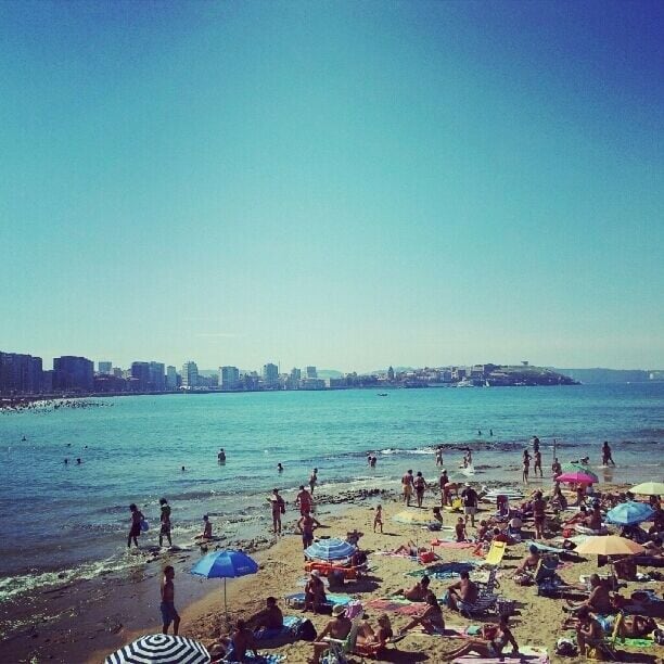 Una playa concurrida donde gente se toma el sol y nada, con sombrillas y un horizonte urbano de fondo.