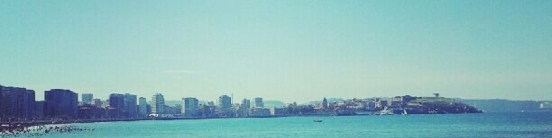 Una playa concurrida donde gente se toma el sol y nada, con sombrillas y un horizonte urbano de fondo.