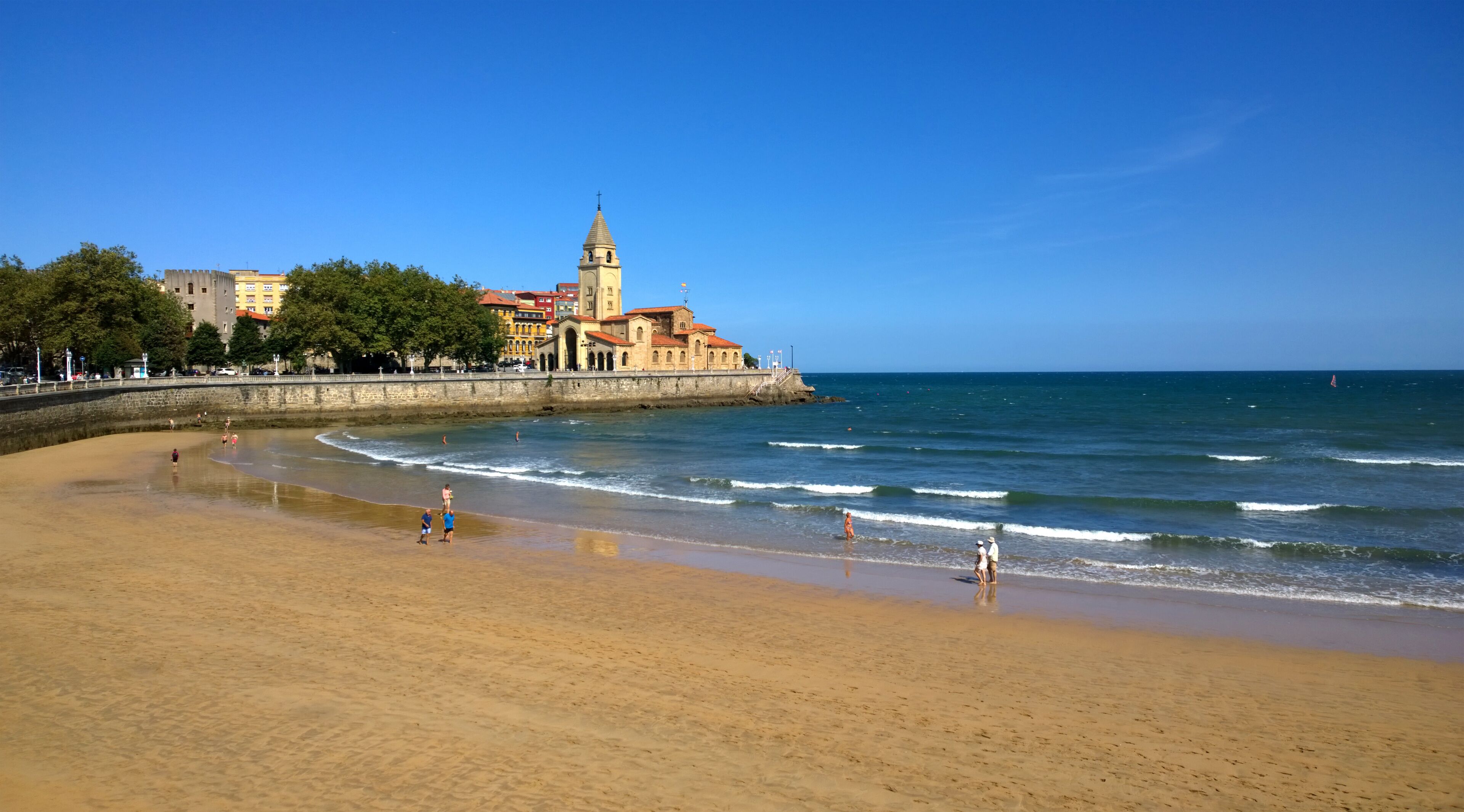 A sandy beach with waves, a church with a bell tower, and a clear blue sky.