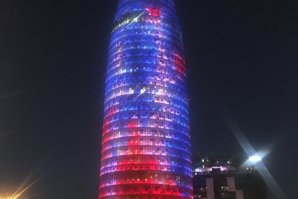 A tall, illuminated tower with a curved design, surrounded by a few people and a staircase leading up to it.