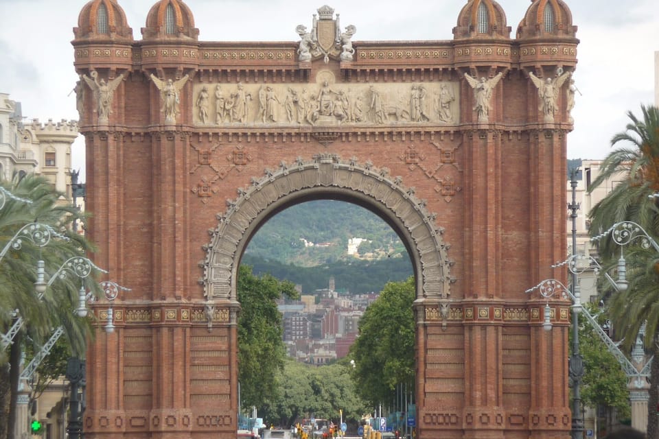 Um grande arco de tijolos vermelhos ornamentados, com esculturas e entalhes elaborados, cercado por palmeiras e um horizonte urbano ao fundo.