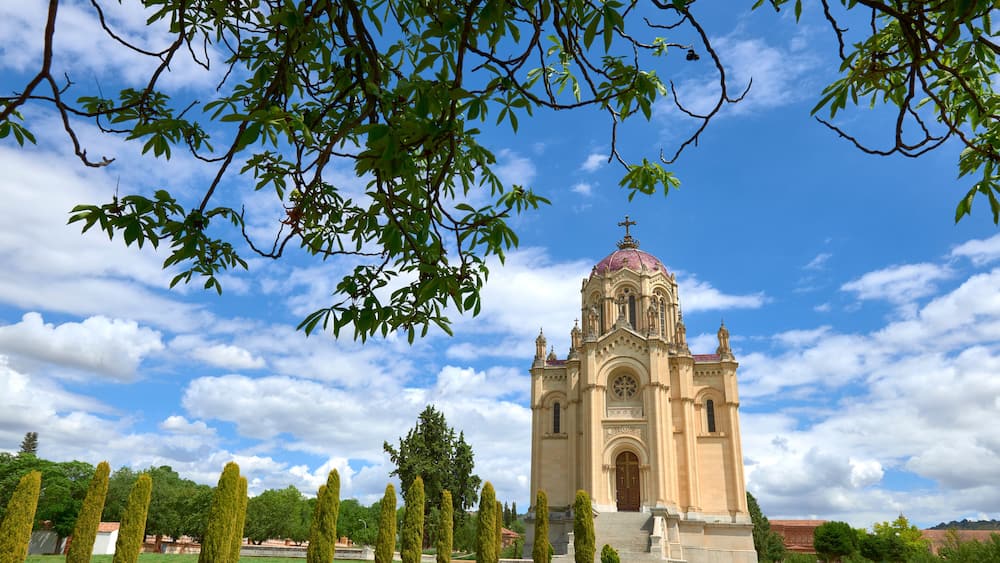 A church with a dome and bell tower, surrounded by neatly trimmed trees and a clear blue sky.