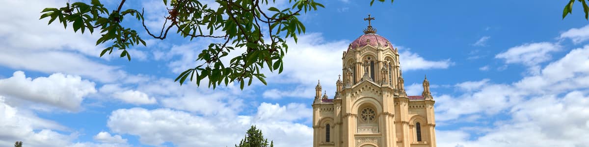 A church with a dome and bell tower, surrounded by neatly trimmed trees and a clear blue sky.