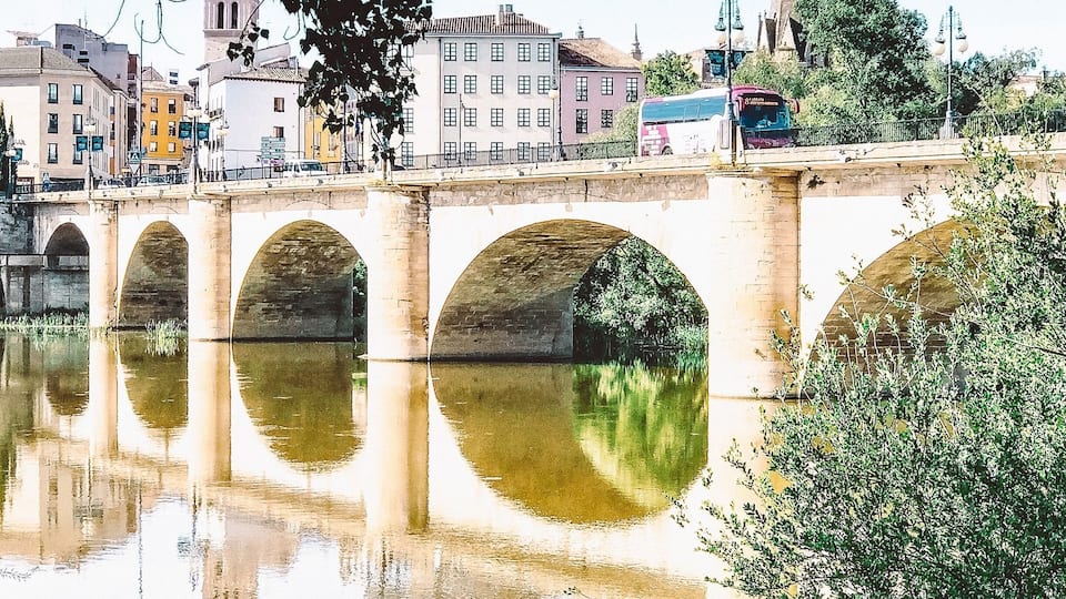 A stone bridge with arches over a calm river, surrounded by greenery and buildings.