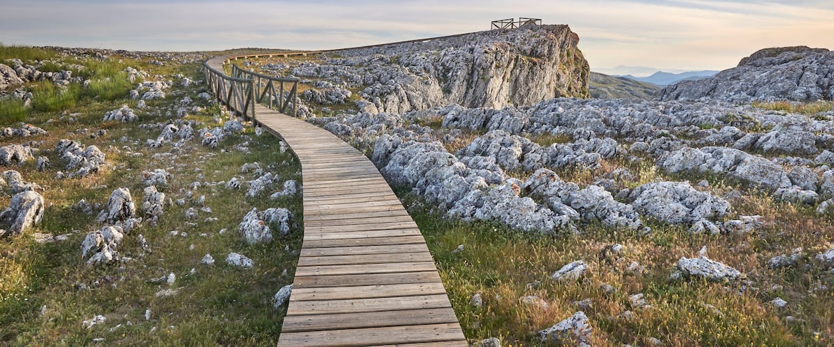 A wooden boardwalk winding through a rocky landscape with a cliff in the distance.