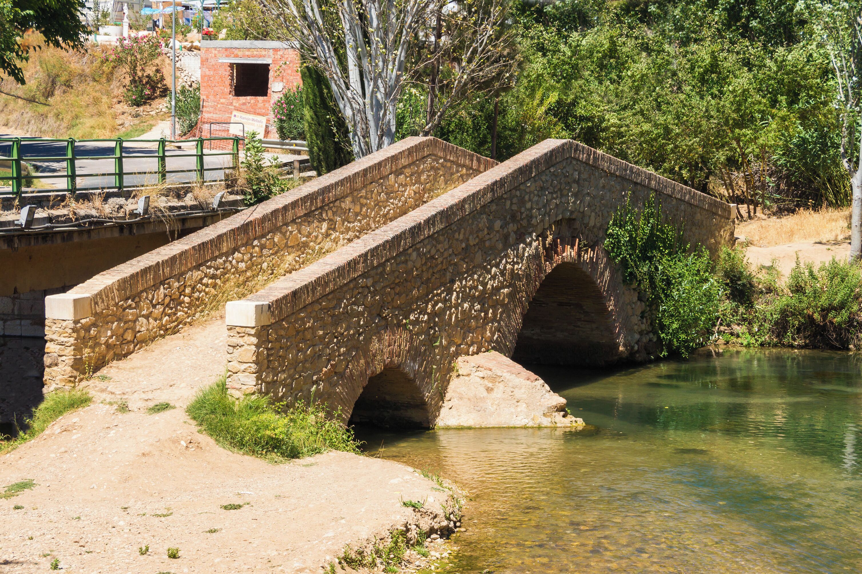 A stone bridge over a river with a dirt path leading to it and a building in the background.