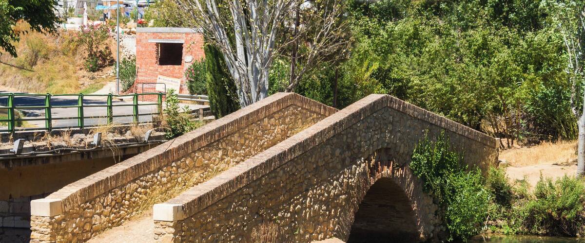 A stone bridge over a river with a dirt path leading to it and a building in the background.