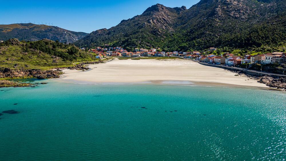 A coastal town with a sandy beach, clear turquoise waters, and mountains in the background.