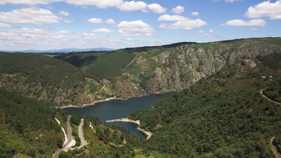 A mountainous landscape with a winding road, a lake, and dense forest.