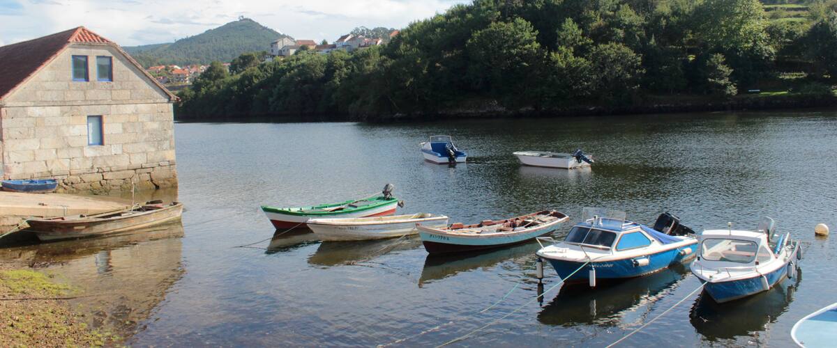A stone building by a calm river with several small boats docked.