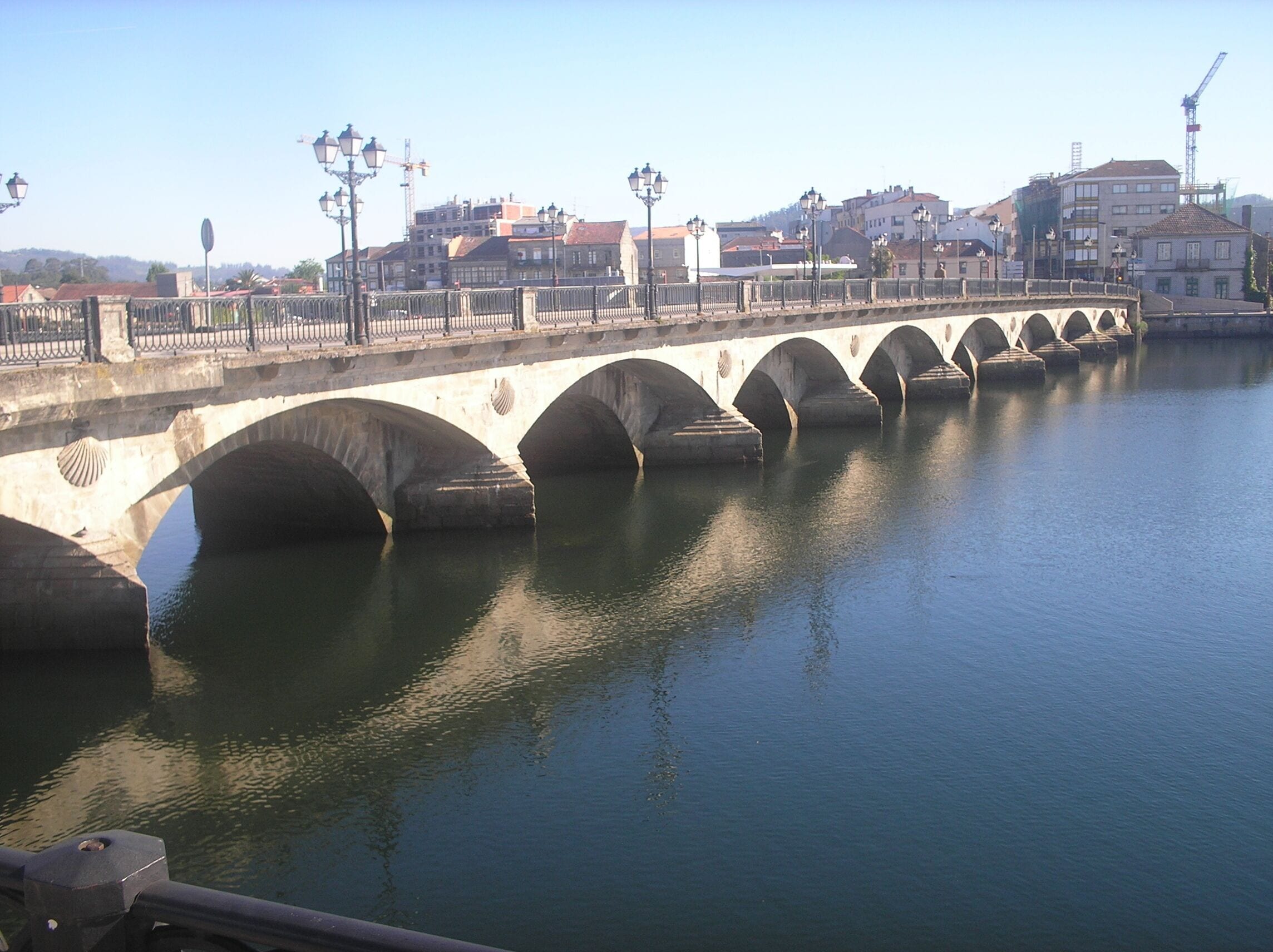 Un puente de piedra sobre un río tranquilo con varios arcos, rodeado de edificios y grúas de construcción.