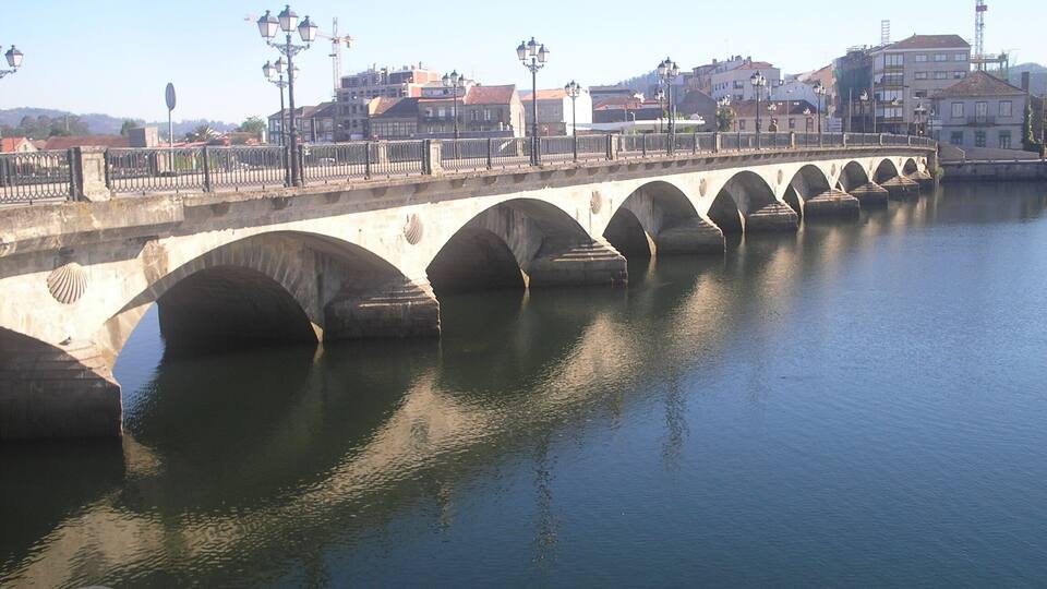 Un puente de piedra sobre un río tranquilo con varios arcos, rodeado de edificios y grúas de construcción.