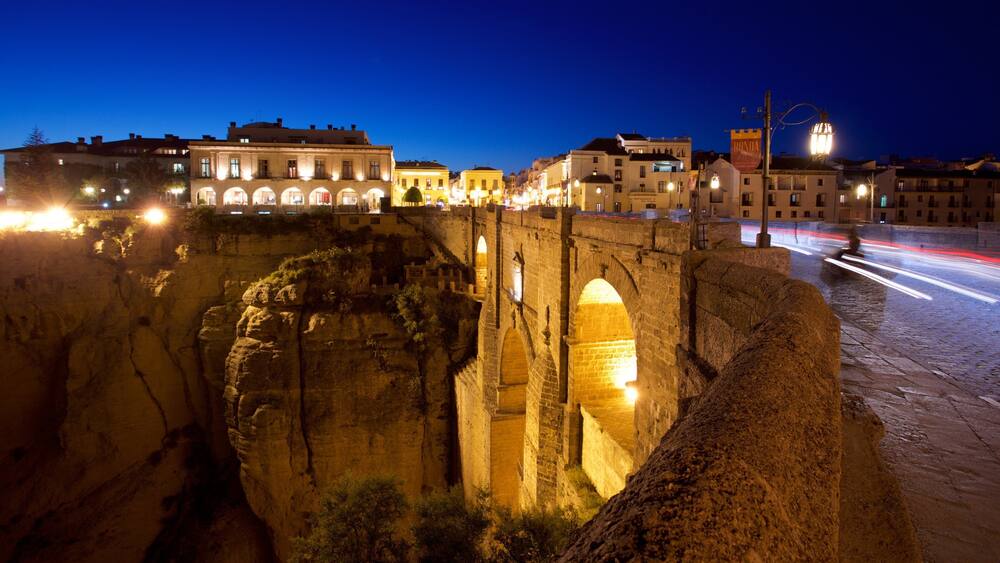 A historic stone bridge at night with illuminated buildings in the background.
