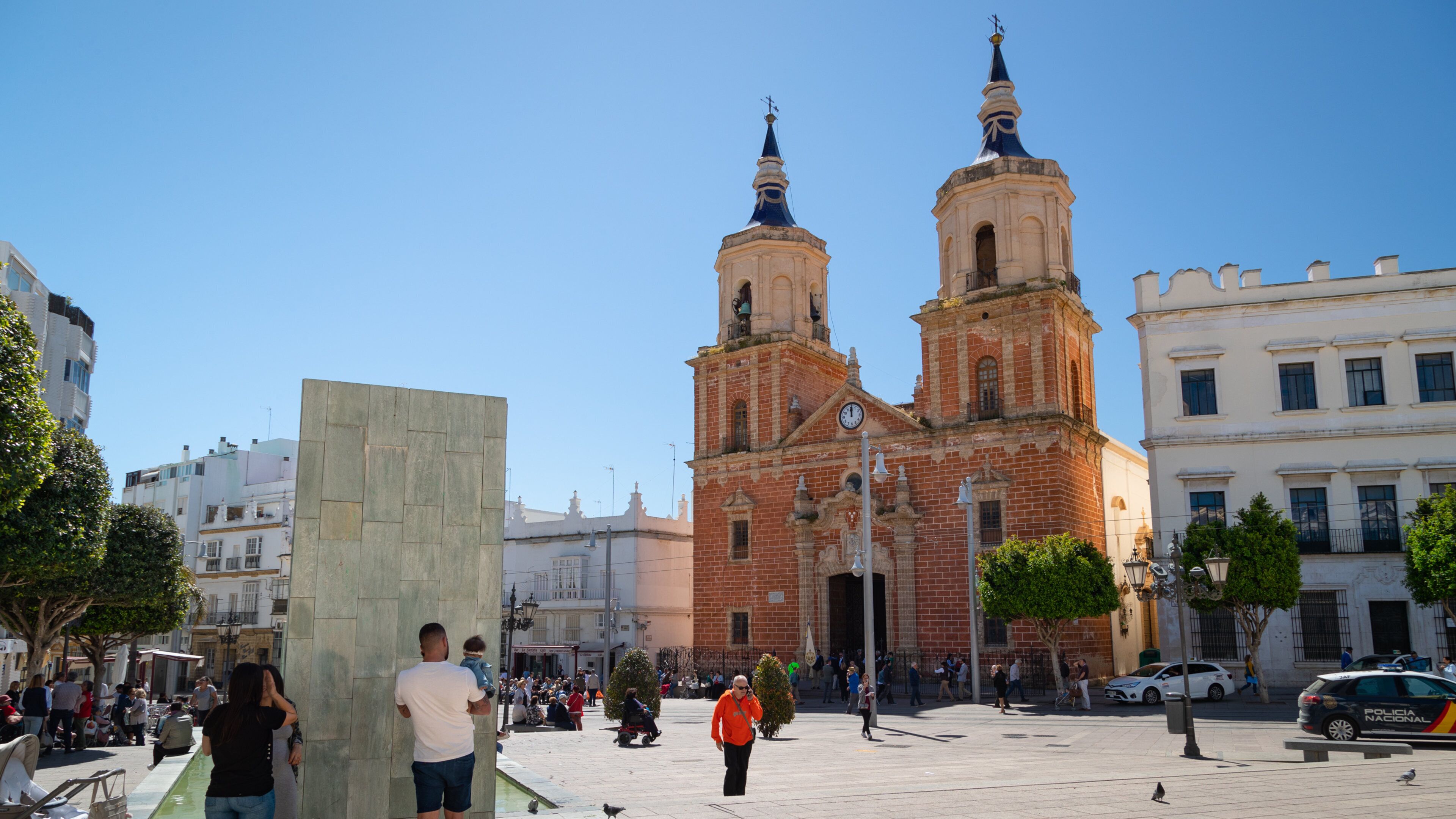 A historic church with two towers, a square with people and a statue, and modern buildings in the background.