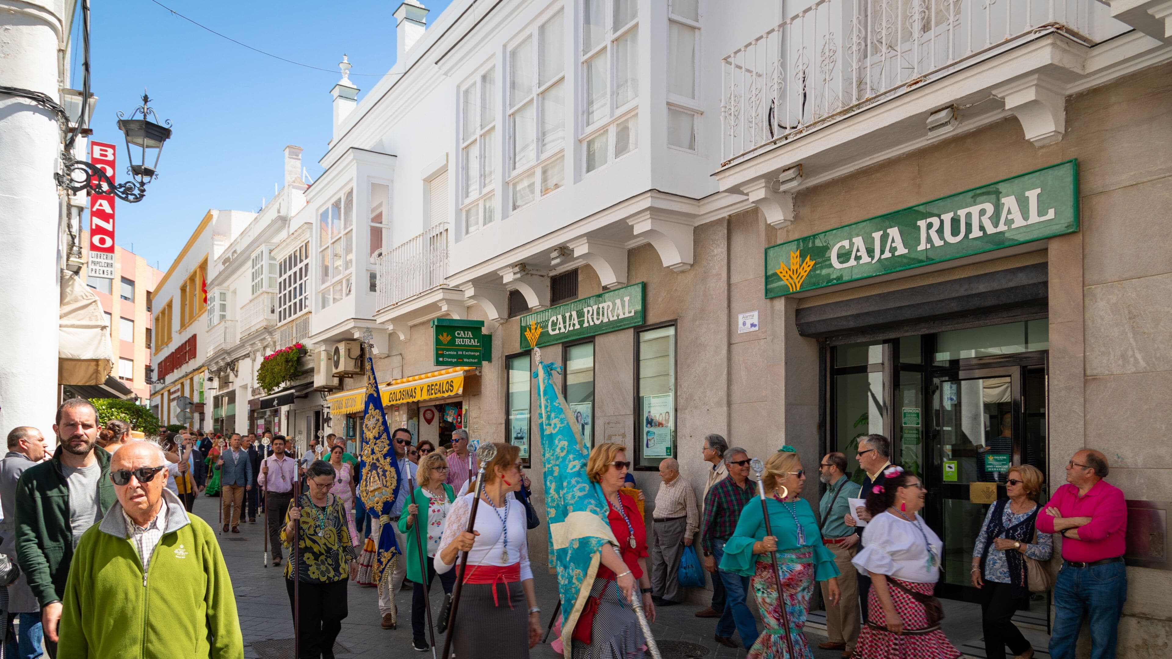 A street with people walking, a building with a sign that reads "CAJA RURAL", and another sign that says "COLOSIMAS Y REGALOS".