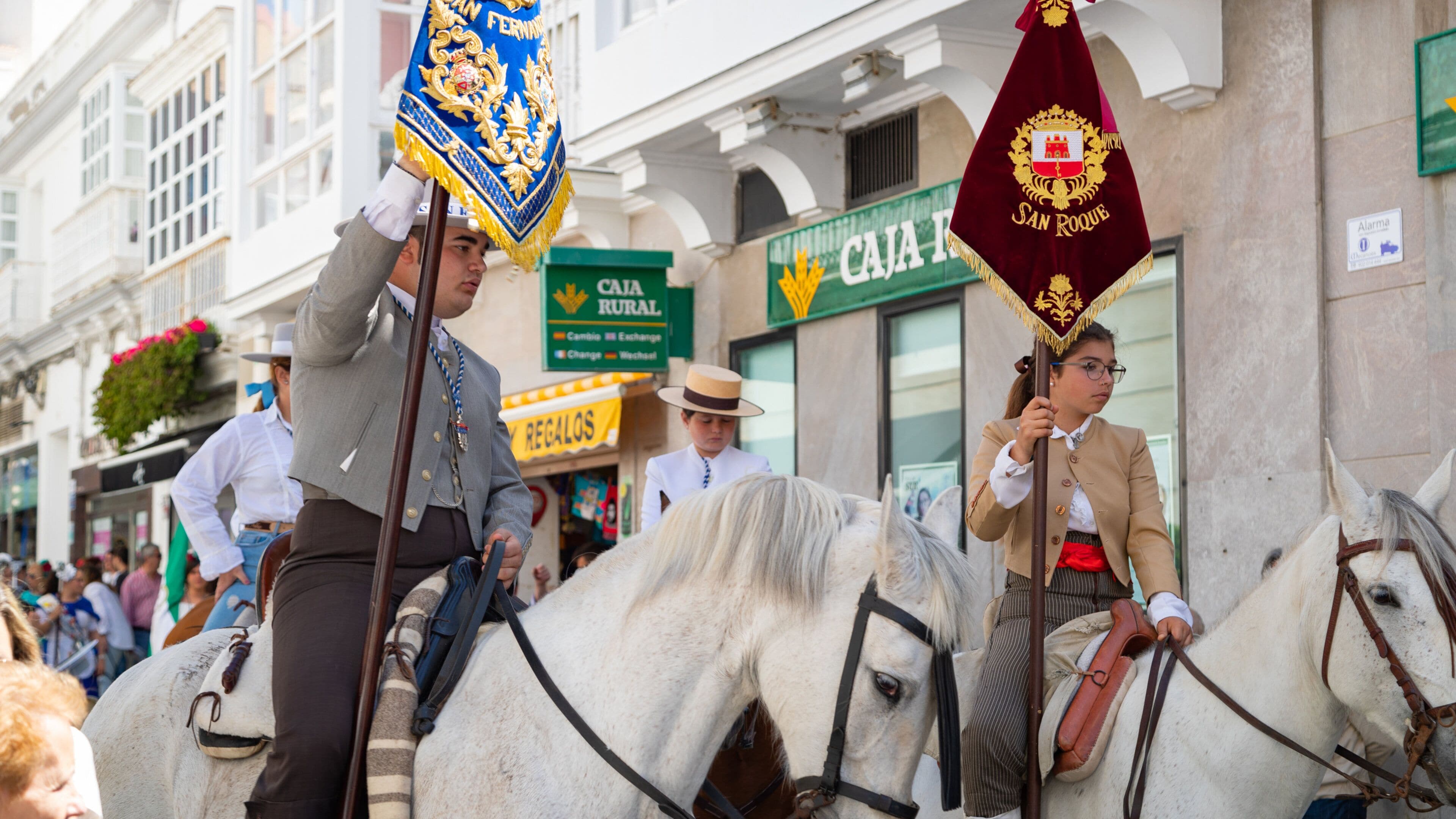 Two individuals on white horses, holding flags, in a street with buildings and a sign that reads "CAJA RURAL".