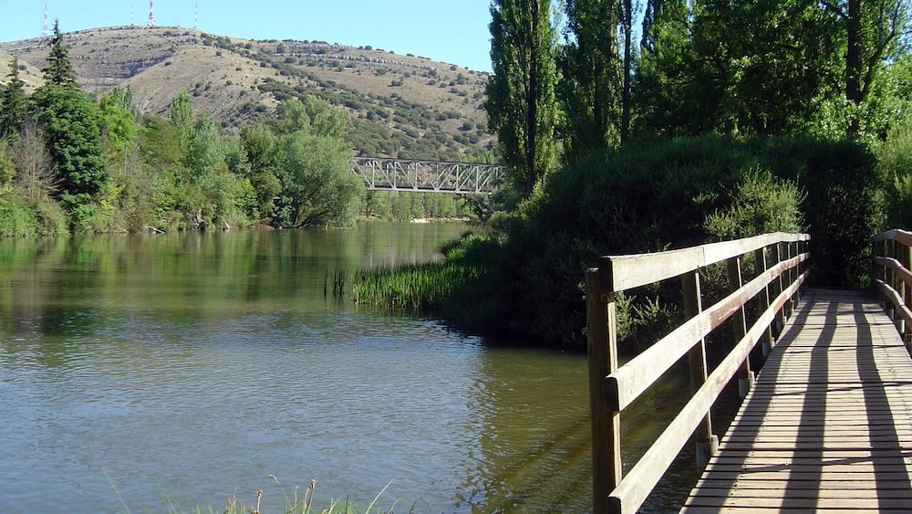 A wooden bridge over a calm river, surrounded by greenery and a distant hill with a metal structure.