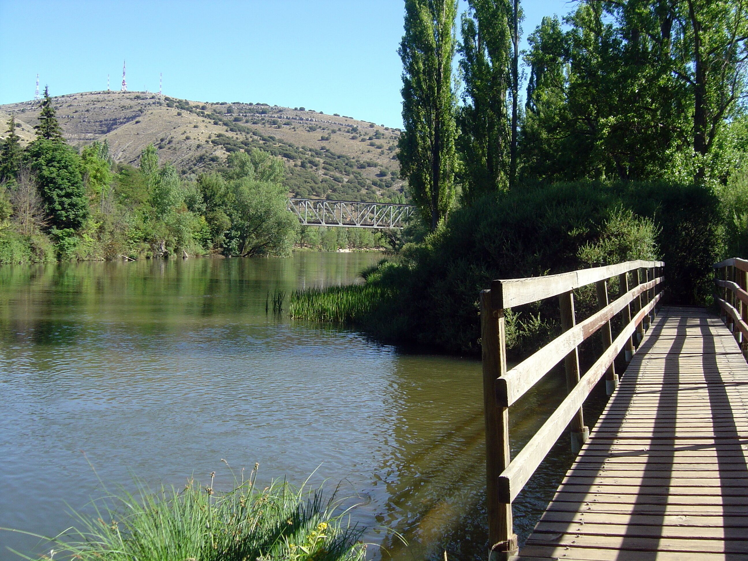 A wooden bridge over a calm river, surrounded by greenery and a distant hill with a metal structure.