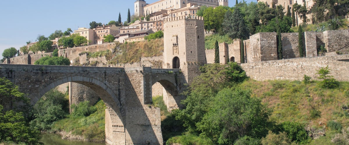 A historic stone bridge over a river, a castle with multiple towers, and lush greenery.