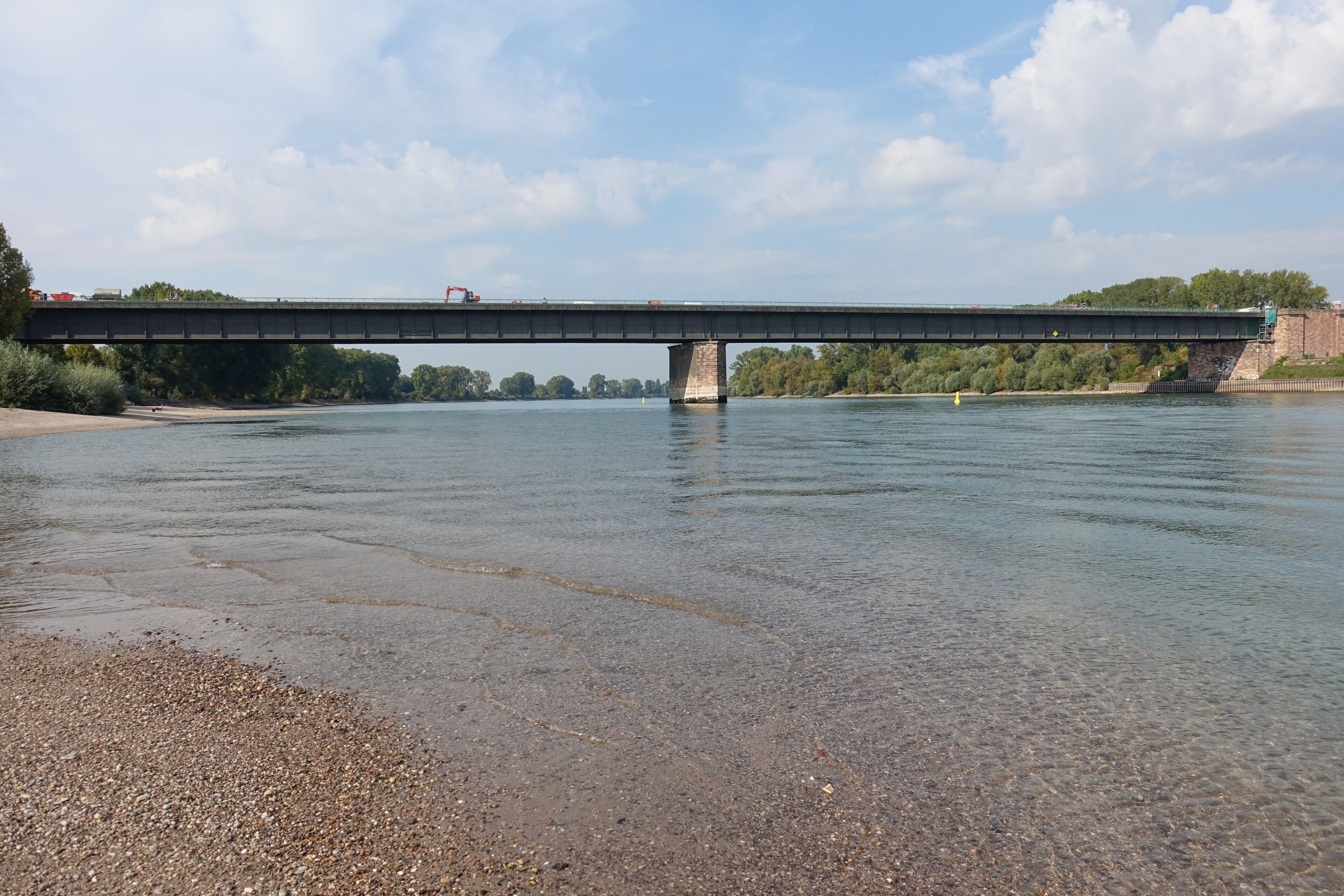 A river with a bridge, pebbly shore, and trees in the background.