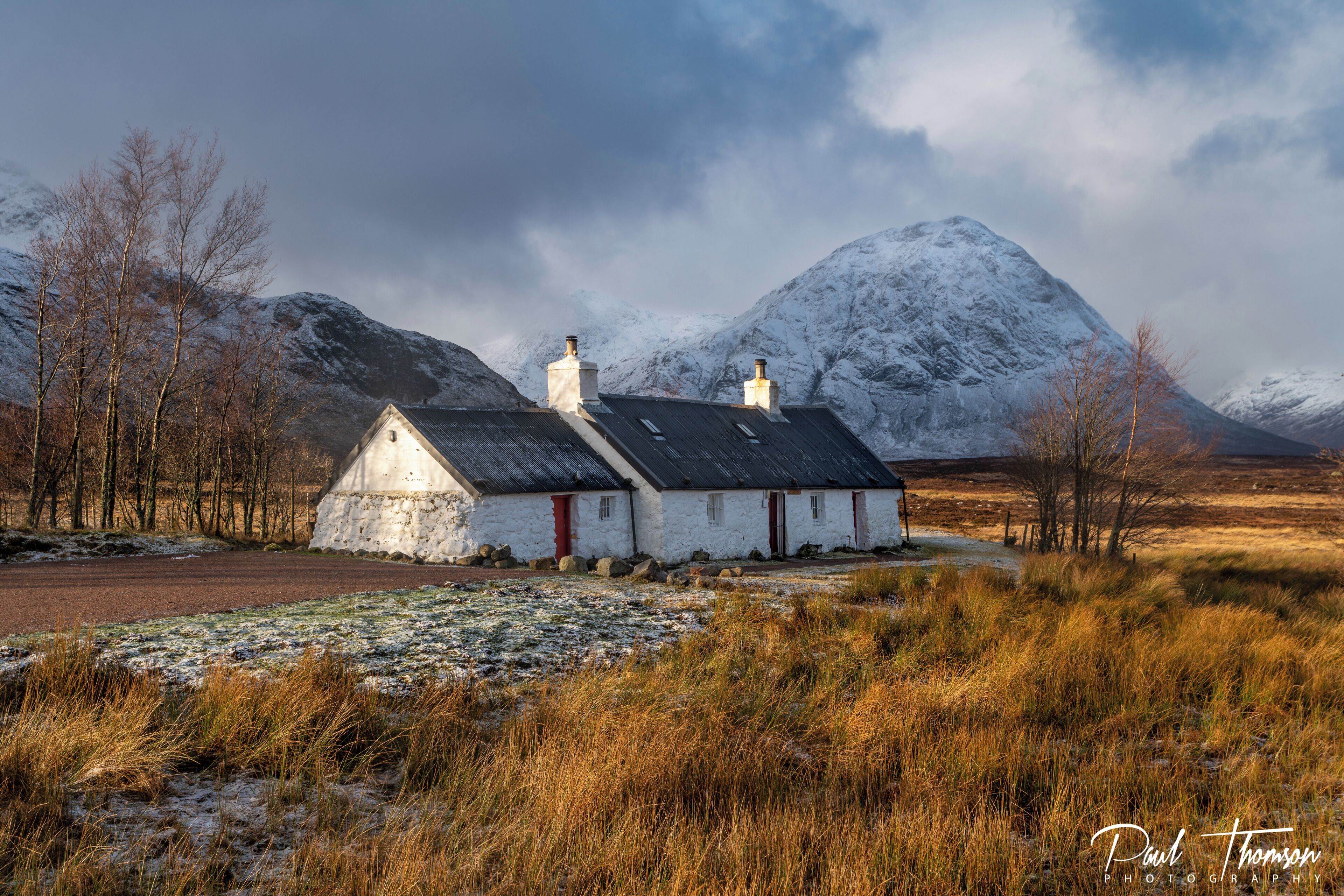 A white house with a red door, surrounded by snow-covered mountains and a grassy field.