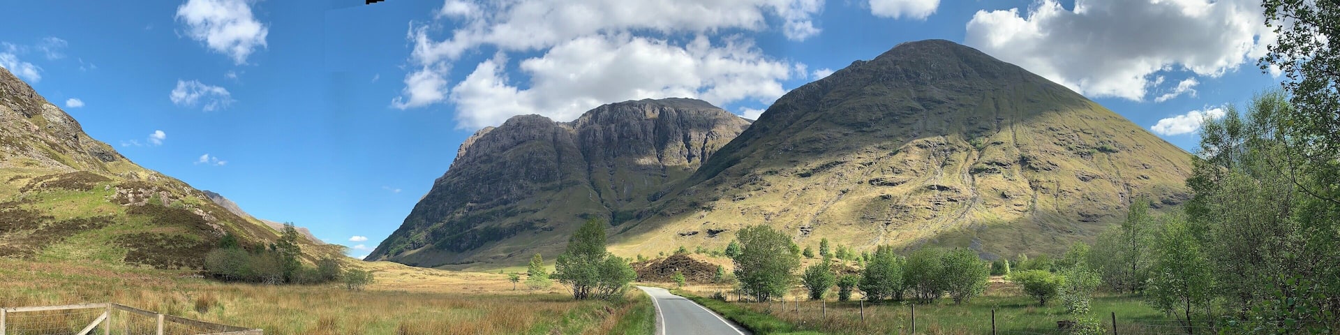 A rural road flanked by grassy fields and mountains under a clear sky.