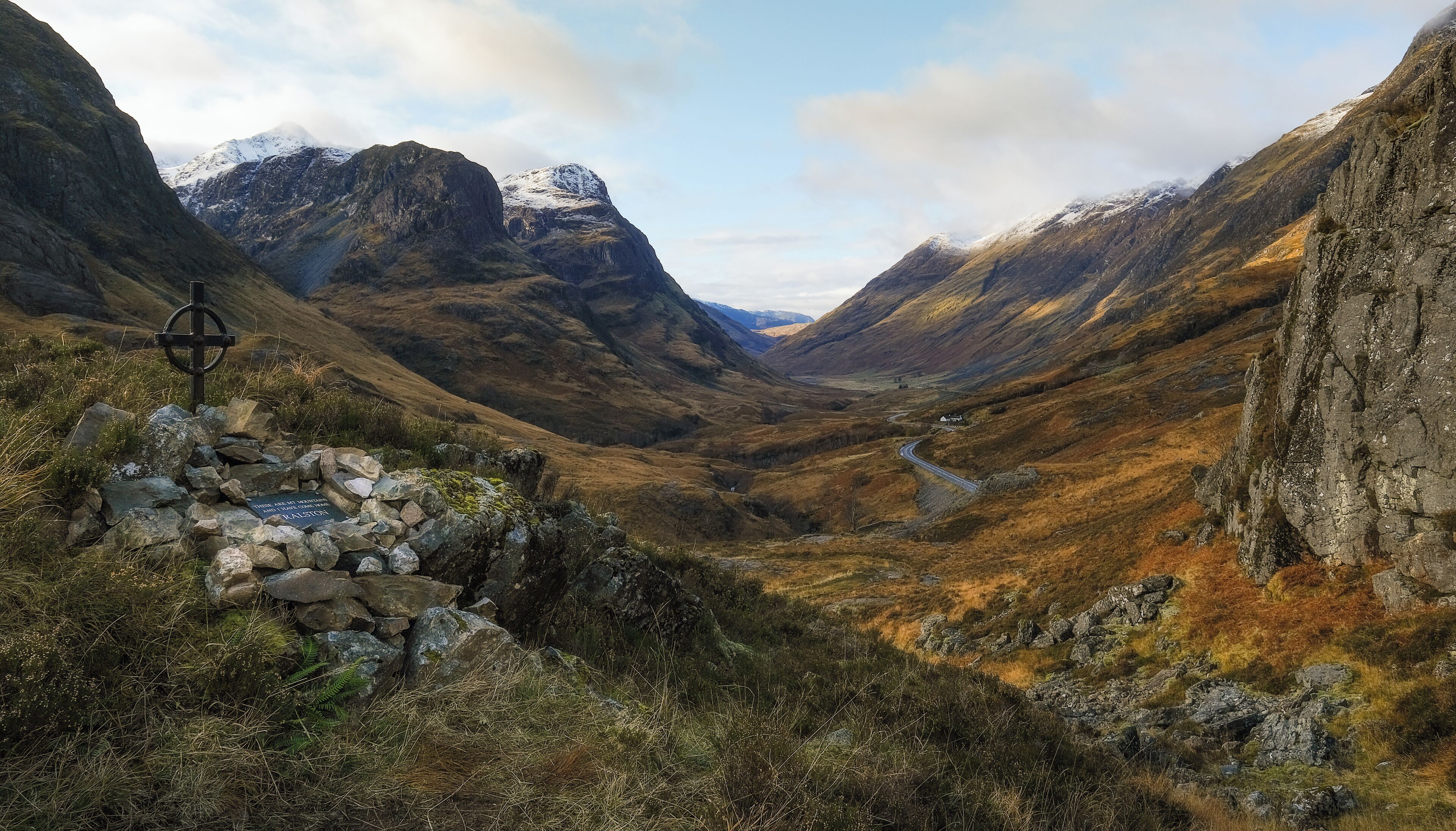 A mountainous landscape with a cairn and a cross, a winding road, and a distant house.