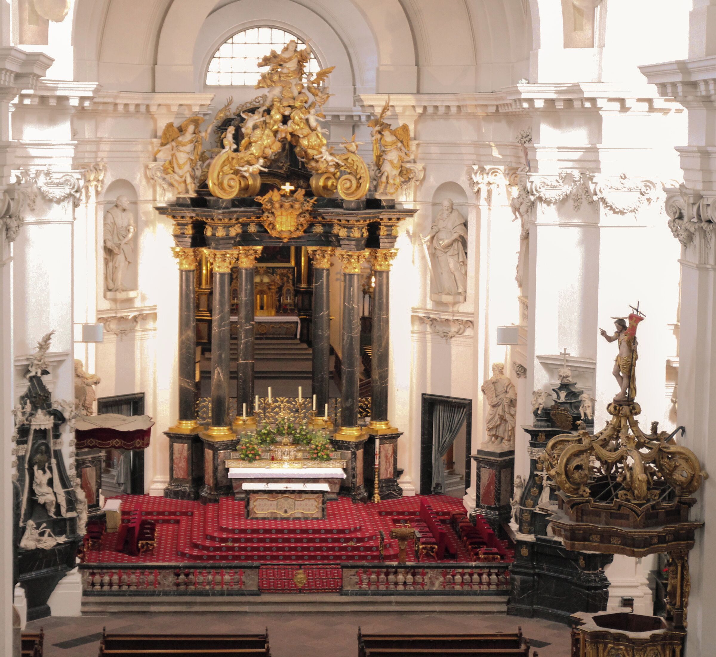 An ornate church interior with a central altar, statues, and red carpeting.