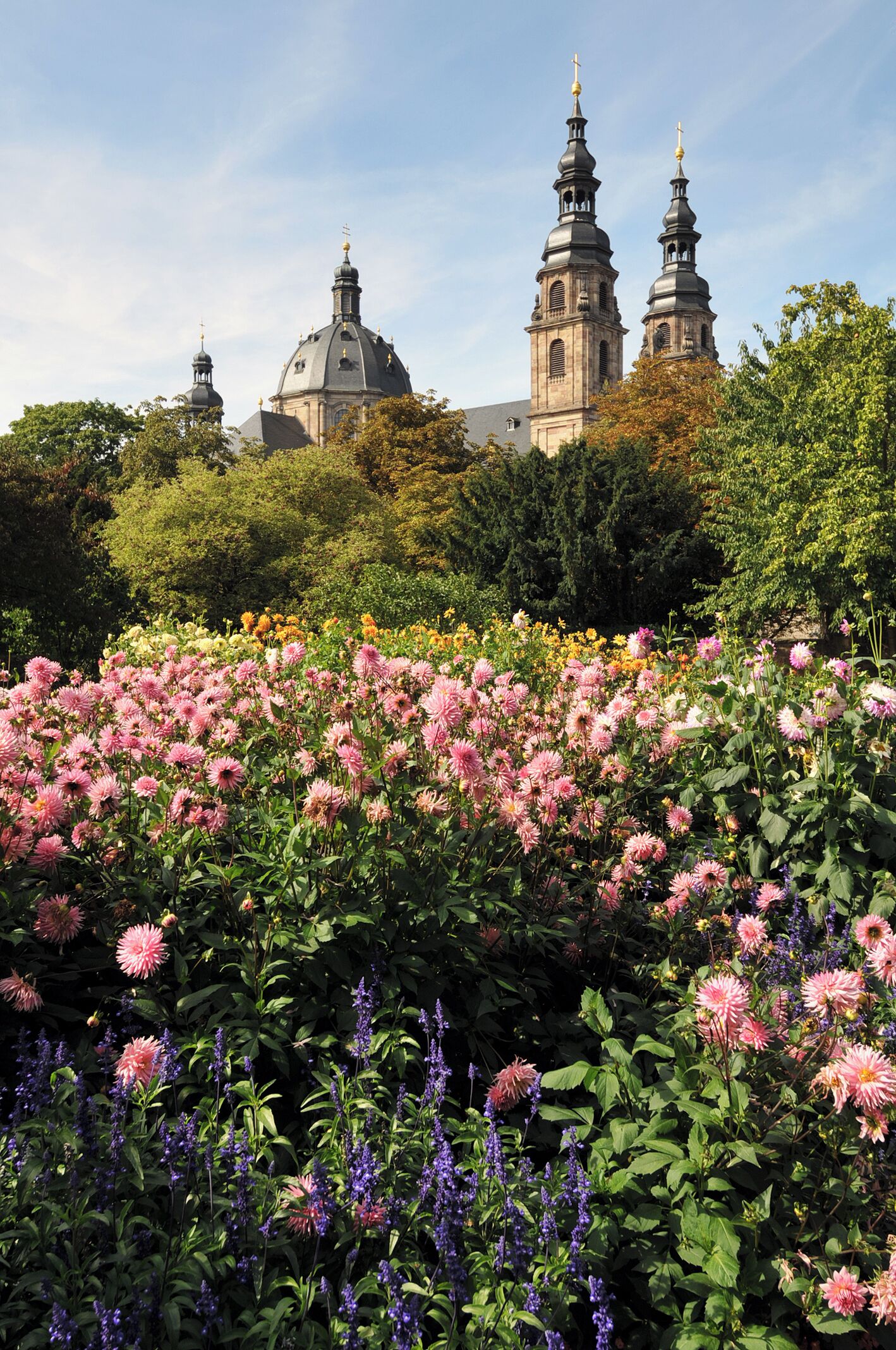 A garden with pink flowers and purple flowers in the foreground, and a church with two towers in the background.
