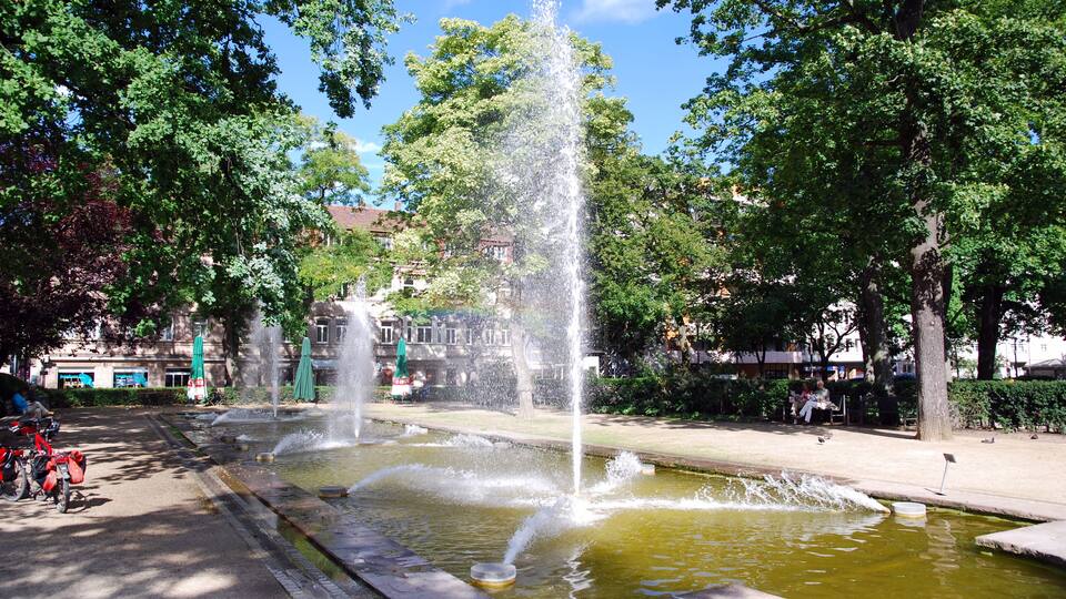 A fountain with water jets in a park with trees and buildings in the background.
