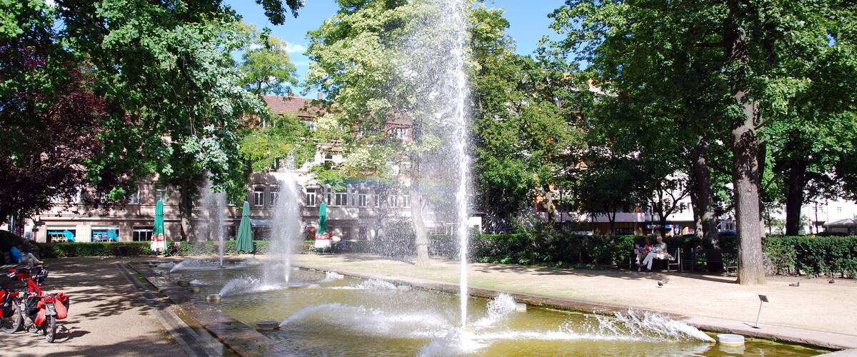 A fountain with water jets in a park with trees and buildings in the background.