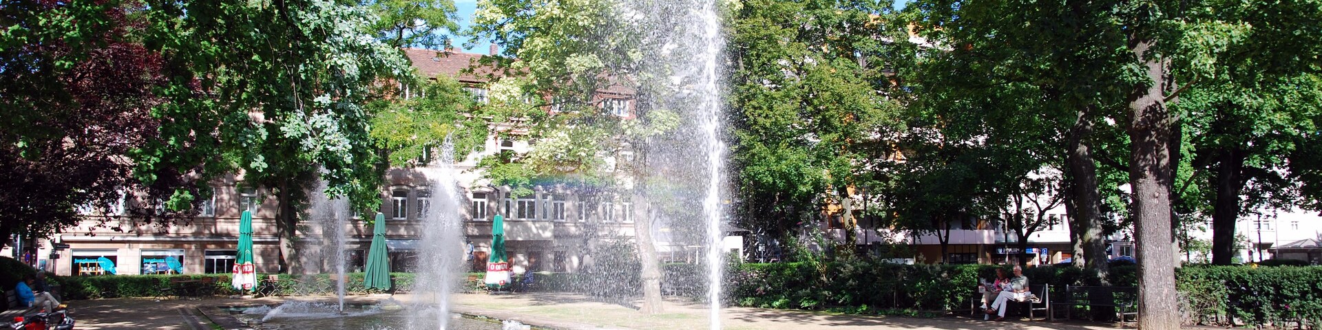 A fountain with water jets in a park with trees and buildings in the background.