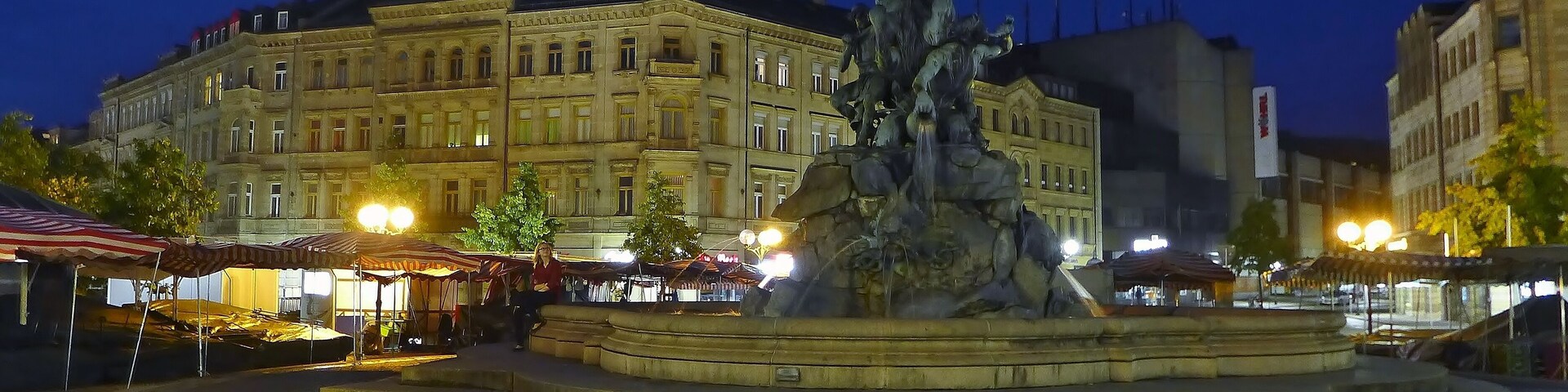 Un edificio histórico de arquitectura clásica, una fuente central con una estatua y faroles de calle circundantes.