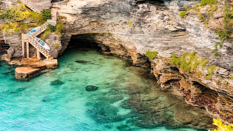 A rocky cave entrance with clear turquoise water, a staircase leading down, and some greenery on the cliff.