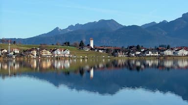 A calm lake with reflections of houses and a church tower, surrounded by mountains.