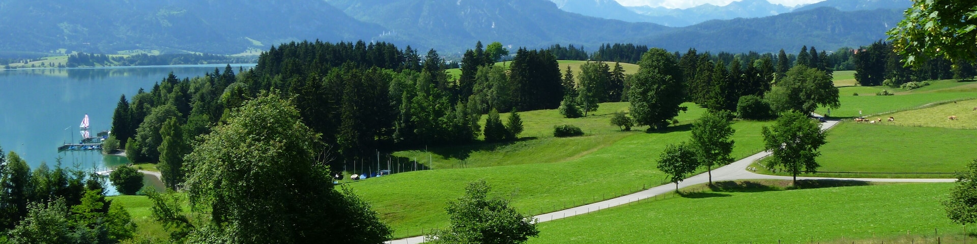 Ein See, umgeben von Bergen, eine Wiese und ein kleiner Steg mit einem Segelboot.