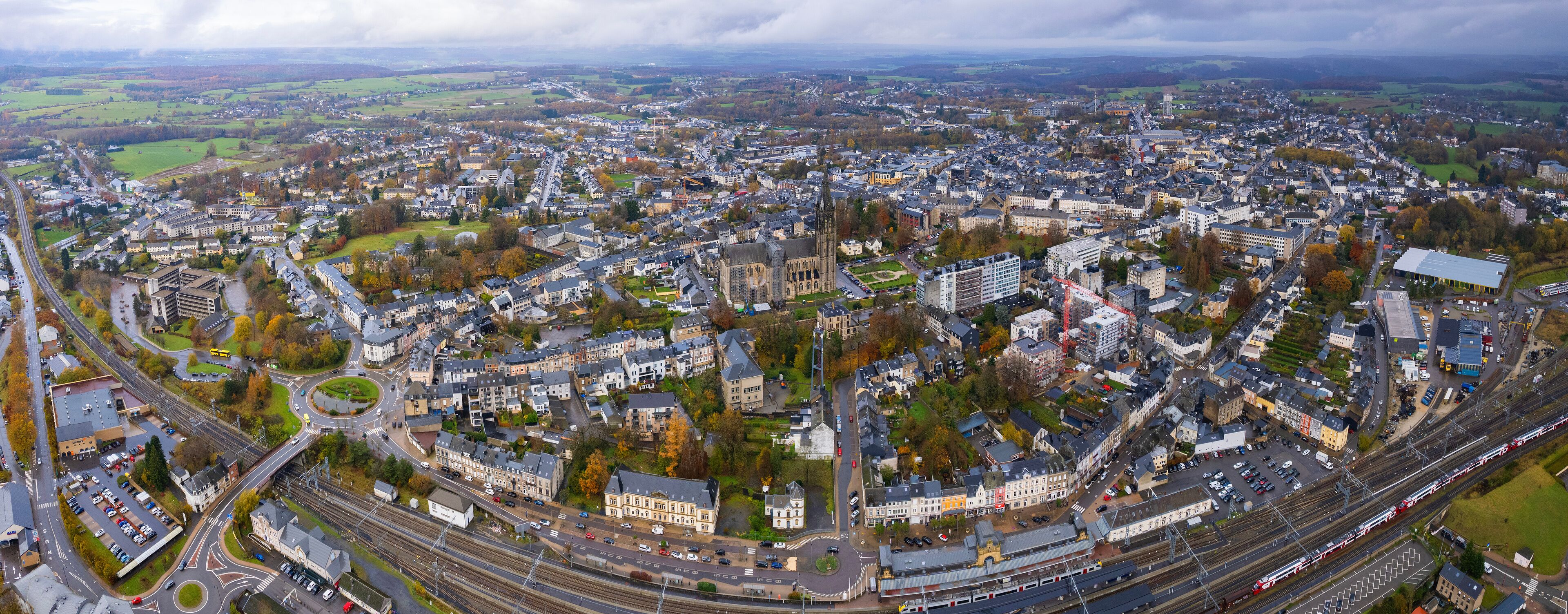 An aerial view of a city with a prominent church, railway lines, and surrounding green fields.
