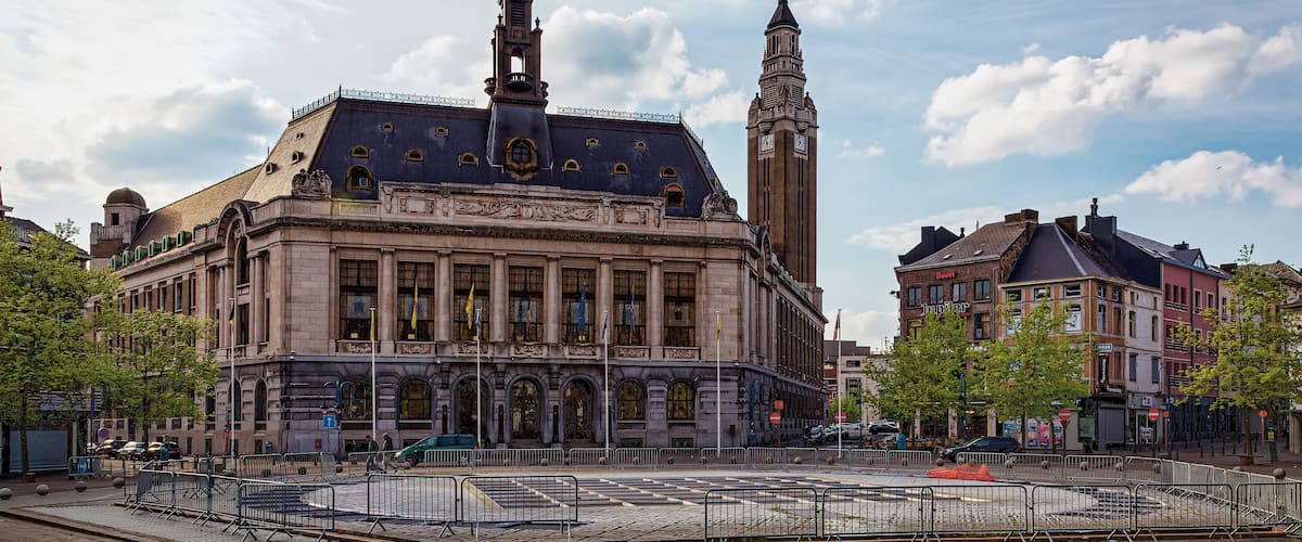 A historic building with a clock tower, surrounded by a plaza with bollards and a fenced area.