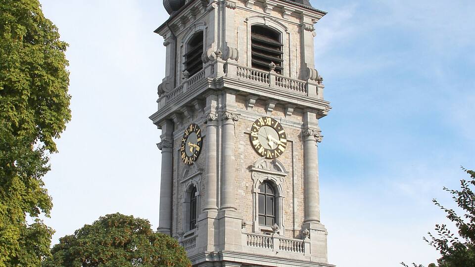 A historic clock tower with multiple domes and a large clock face, surrounded by lush green trees.