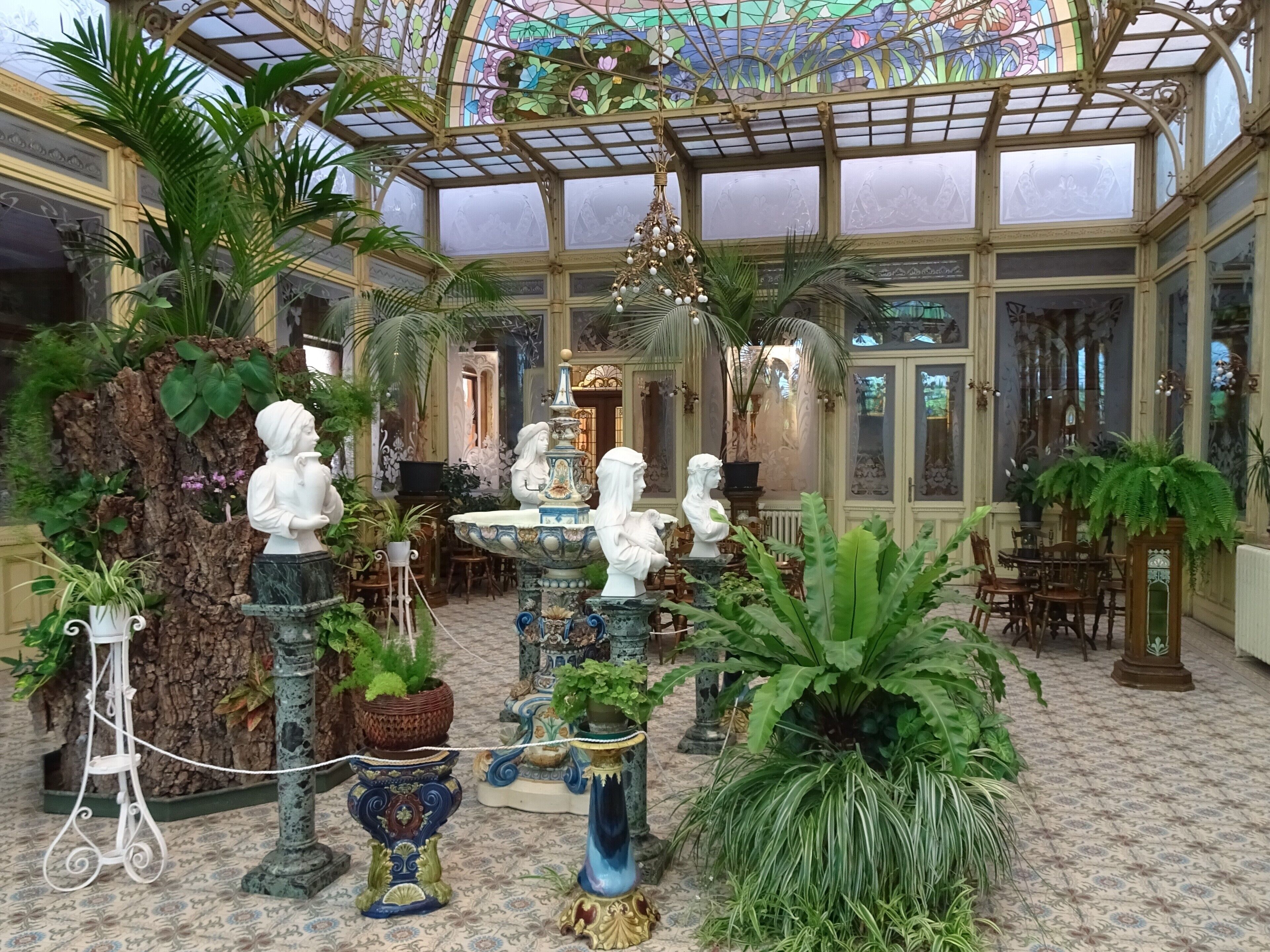 A conservatory with ornate glass ceiling, statues, and lush greenery.