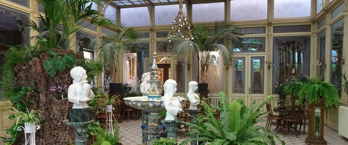 A conservatory with ornate glass ceiling, statues, and lush greenery.