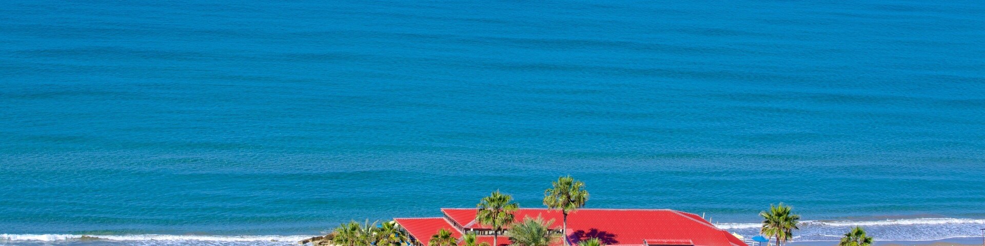 Un resort frente al mar con un edificio de techo rojo, palmeras y un océano azul cristalino.