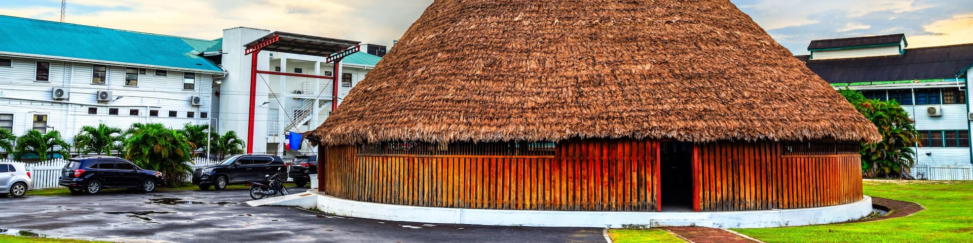 A thatched-roof hut with a white exterior, surrounded by a paved area and a building with a blue roof in the background.