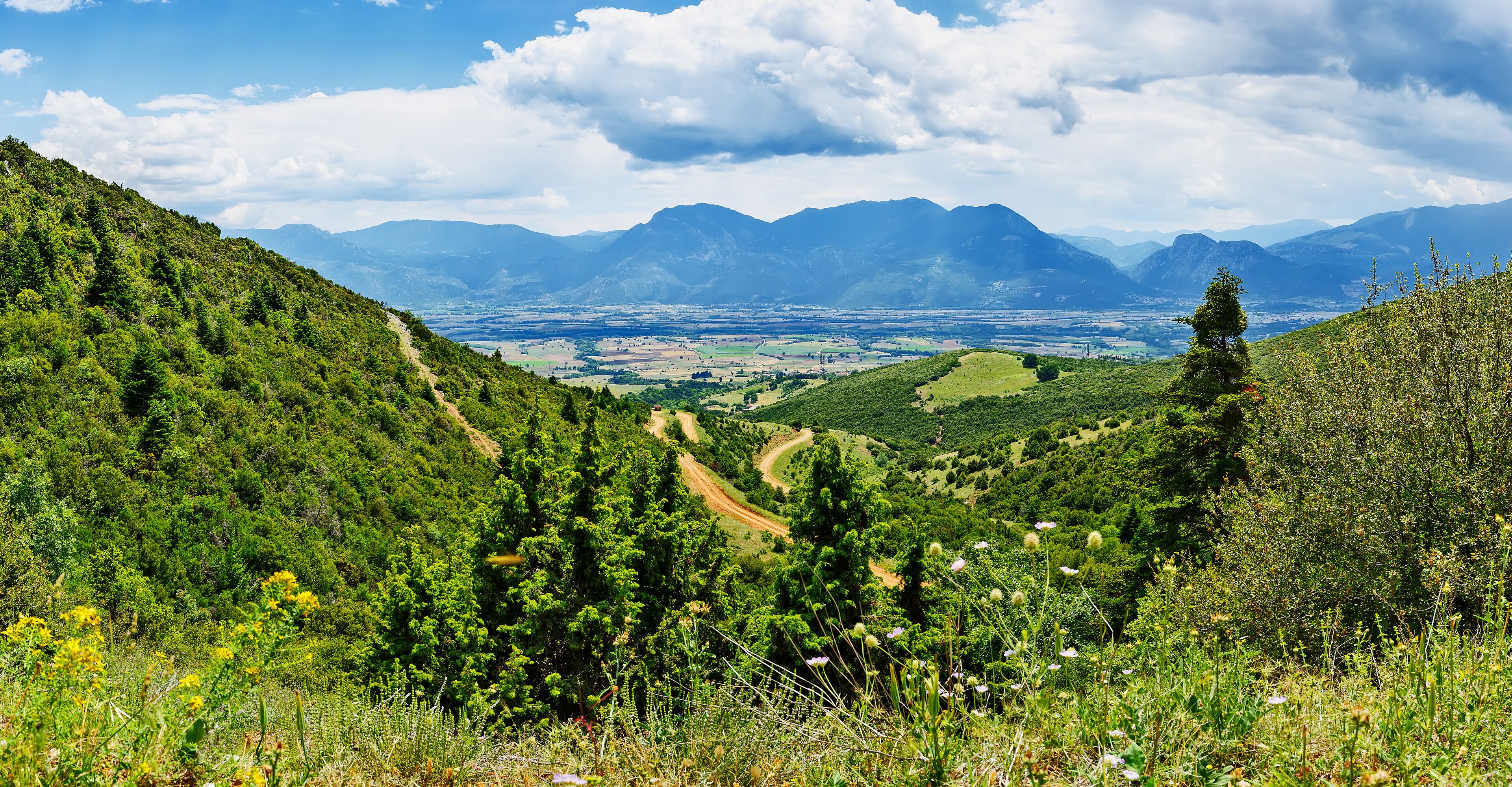 A mountainous landscape with a valley, a dirt path, and distant mountains under a partly cloudy sky.