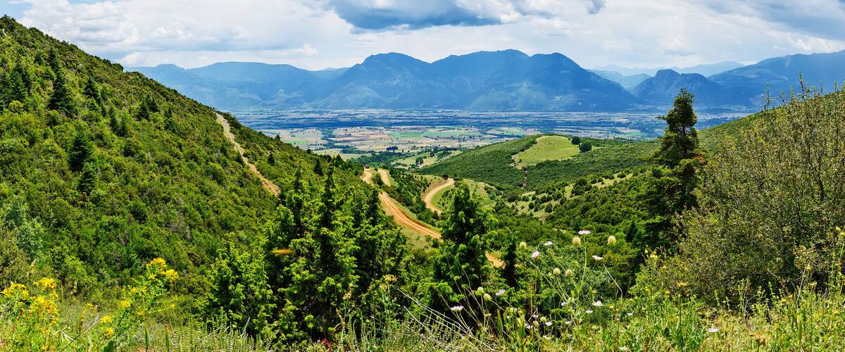 A mountainous landscape with a valley, a dirt path, and distant mountains under a partly cloudy sky.