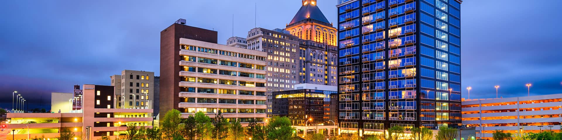 A cityscape at dusk with illuminated buildings, a parking garage, and a landscaped area with trees.