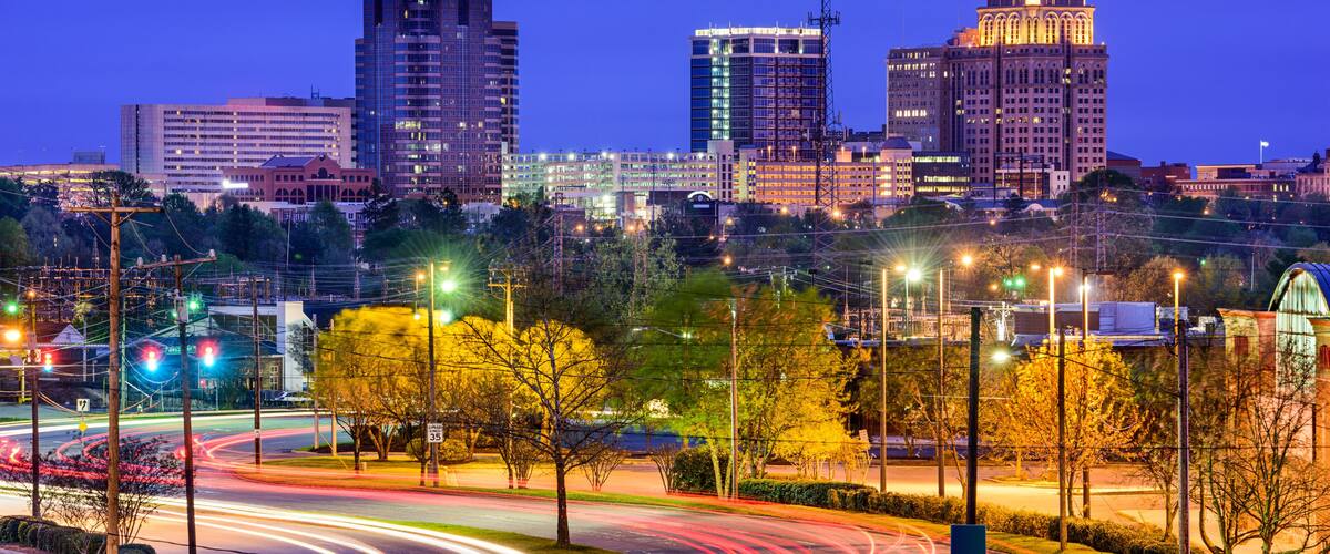 A city skyline at dusk with illuminated buildings, a roundabout with light trails, and a sign indicating 'SOUTH 220'.